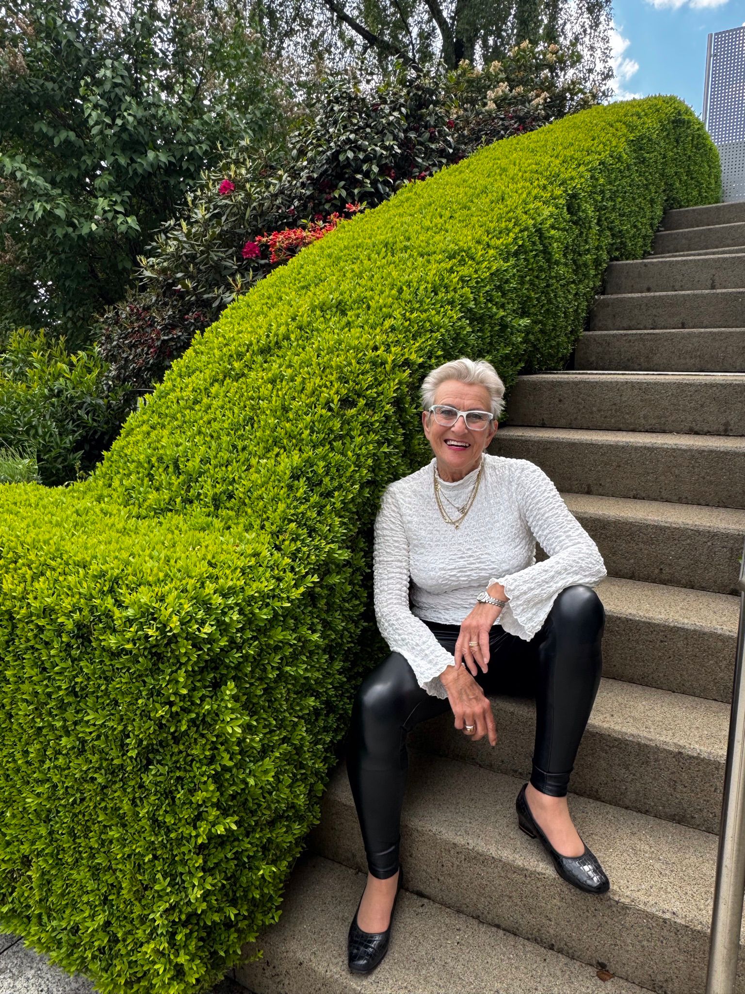 Frau auf Treppe Auf dem Bild sieht man eine ältere Dame mit einem weißen Pullover und einer schwarzen Hose auf einer Treppe sitzen. An ihrer Schulter sieht man eine grüne Hecke.