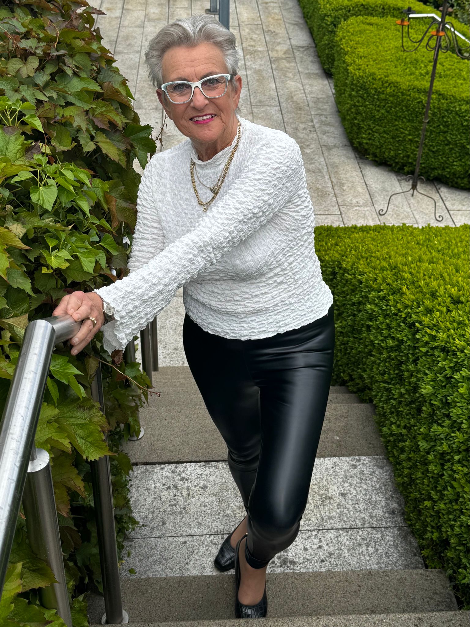 WhatsApp Image 2025-08-11 at 14.48.25 Ältere Dame läuft in die Kamera lächelnd eine Treppe hoch und berührt mit beiden Händen ein Geländer, sie trägt einen weißen Pulli und eine schwarze Hose, man sieht eine grüne Hecke an ihrer Seite und eine graue Terasse im Hintergrund. Die Dame trägt eine Brille.