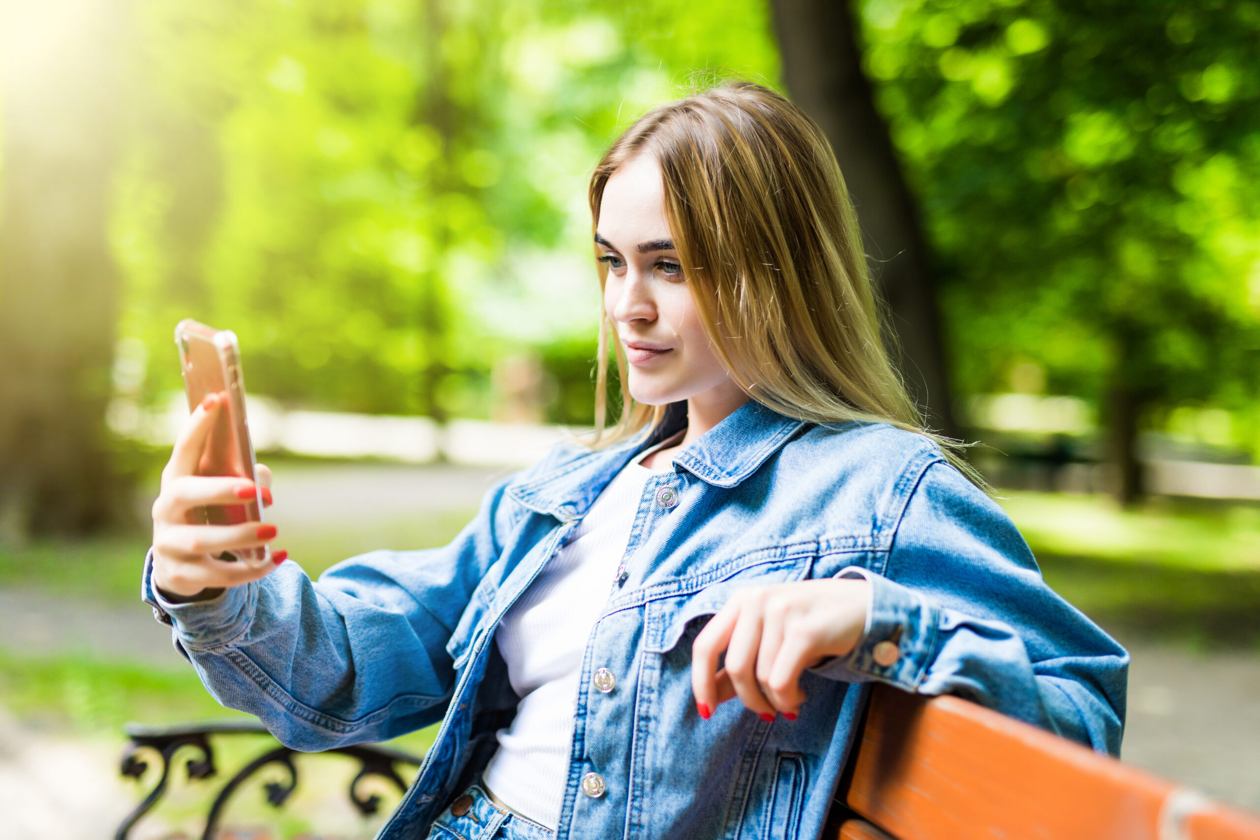 Happy girl using a phone in a city park sitting on a bench Blonde Frau sitzt im Park auf einer Bank und macht ein Selfie mit einem Handy. Sie trägt eine blaue Jeansjacke. Die Bank ist orange.