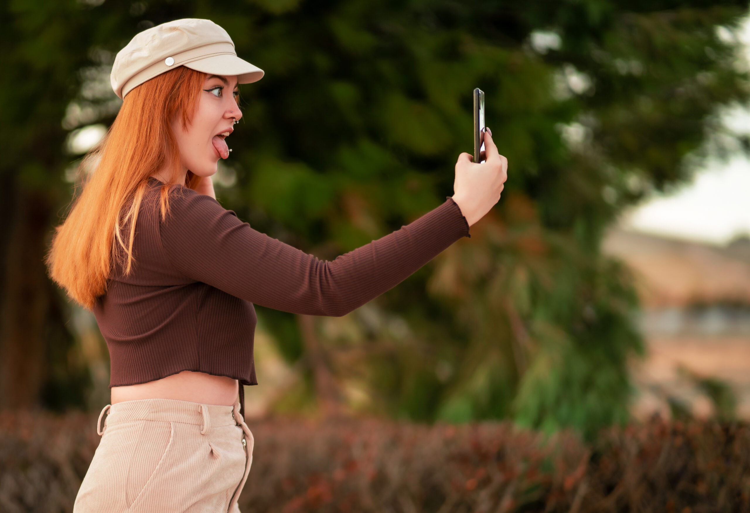Young Caucasian red-haired woman doing a selfie with a funny face Junge Frau mit roten, längeren Haaren und einer beigen Kappe macht ein Selfie mit einem lustigen Gesicht. Dabei streckt sie die Zunge raus.