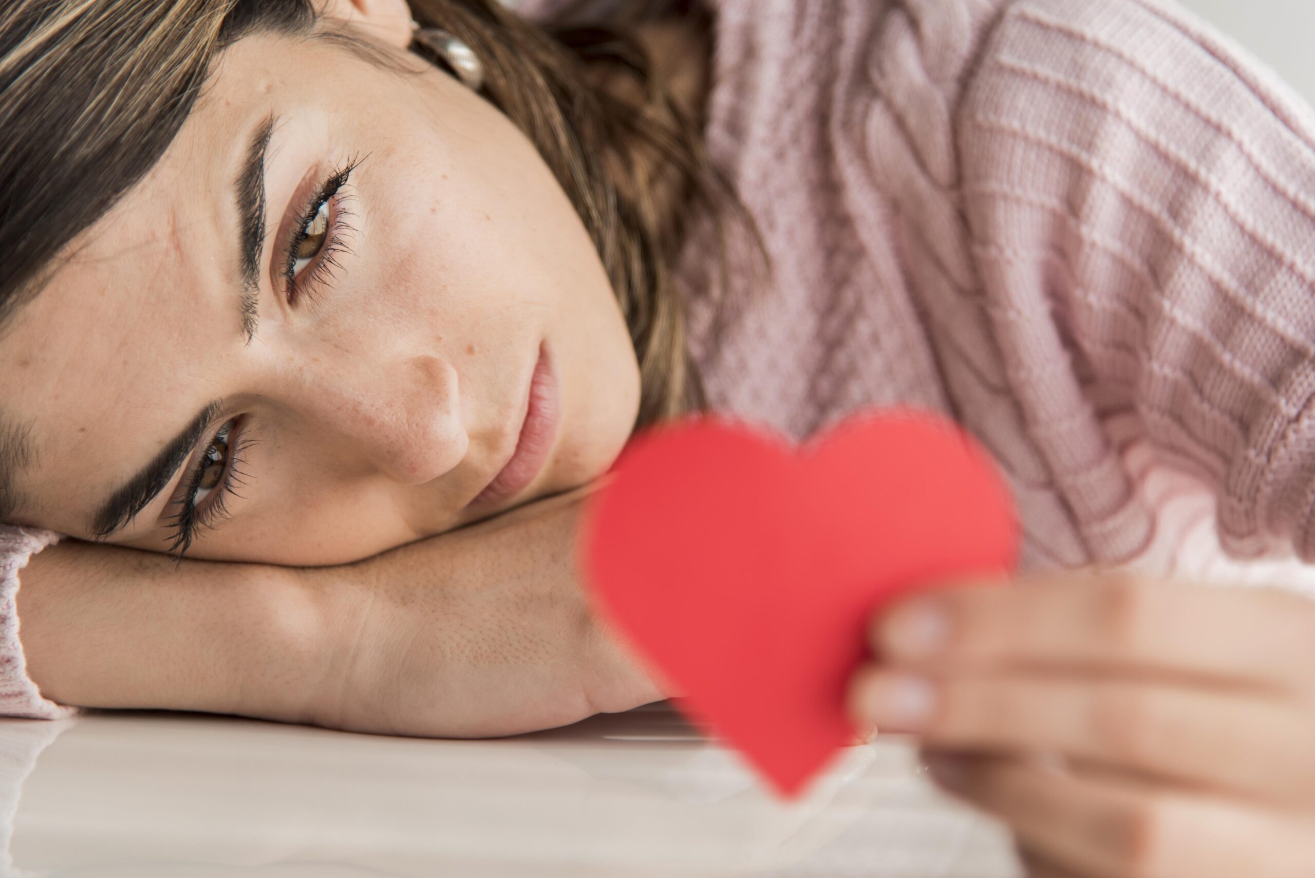 nahaufnahmefrau-die-herz-halt Liegende Frau, Nahaufnahme von ihrem Gesicht, sie liegt auf ihrer Hand und hält ein rotes Herz in der anderen Hand, welches sie verträumt anschaut. Die Frau ist brünett.